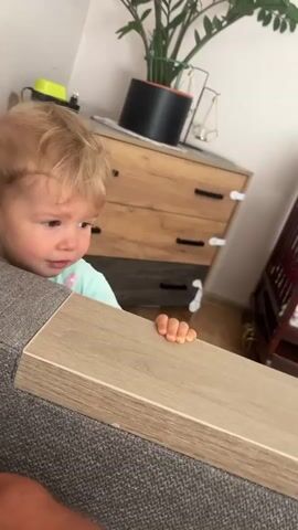 A young toddler with blonde hair and brown eyes is leaning over a wooden surface. The toddler is wearing a light blue t-shirt with a pink floral pattern. The toddler's hands are gripping the edge of the wooden surface. In the background, there is a dresser with drawers, a plant in a black pot, and a crib. The room appears to be a bedroom or living area. The toddler is making facial expressions, suggesting they may be distressed or about to cry.