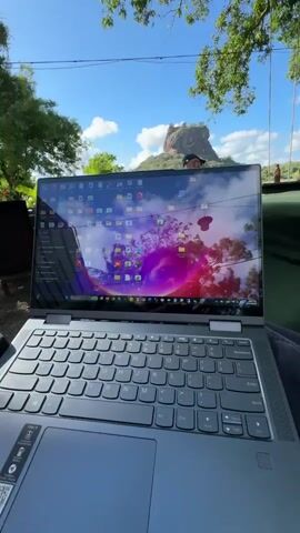 Here is a description of the video:
The video shows a view of Sigiriya rock in Sri Lanka from an outdoor restaurant. A laptop sits in the foreground, and a man is seated at a table nearby. The area appears to be a garden, with trees providing shade over several tables, and a gravel surface. The sky is blue with scattered clouds.