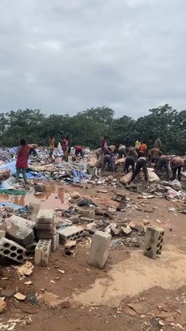 The video shows a group of approximately fifteen people, mostly men and young males, engaged in the manual sorting and removal of debris from a large, open-air dump site. The ground is muddy and uneven, covered in a wide assortment of refuse including broken concrete blocks, plastic sheeting, fabric scraps, and other unidentifiable waste materials. The individuals are dressed in casual, everyday clothing, with some wearing shirts in red, orange, and green. They are actively sifting through the piles of trash, some carrying large bags, suggesting they are collecting specific items or clearing the area. The background consists of lush green trees under an overcast sky. The overall scene depicts a labor-intensive clean-up operation in a significant waste disposal area.