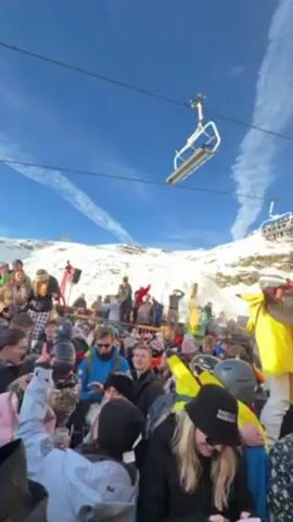 A video shows a lively après-ski party at a mountainside bar, likely in Europe. A large crowd of people dressed in ski gear and casual clothing are gathered outside a wooden building labeled "FOLIE’S SHOP." Many people are dancing and singing along to music, appearing to have a good time. The crowd is dense, with faces clearly visible. Some individuals are wearing sunglasses, ski helmets, and colorful jackets. The bar structure is two stories tall, with speakers mounted on the exterior. A ski lift runs above the scene, with a few empty chairs in view. The background shows a snow-covered mountain under a clear blue sky with contrails. The overall atmosphere is festive and energetic.