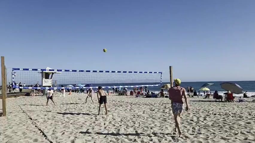 A group of people are playing a game of volleyball on a sandy beach on a clear, sunny day. The ocean is visible in the background, along with several beach umbrellas and people lounging in the sand. A lifeguard stand is also present on the left side of the frame. The video captures a moment of action during the game, with players in motion, the ball in the air, and others watching from the sidelines.