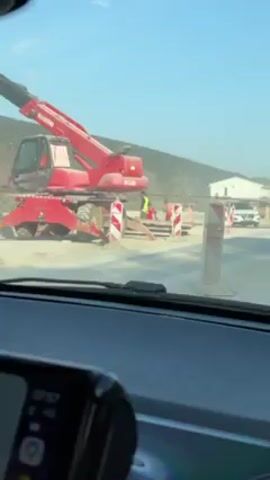 The video shows a view from inside a car, driving through a road construction site. A red Manitou construction vehicle is visible to the left, along with construction workers wearing safety vests and red suits. Traffic cones and signs, including one with "BEMAX" written on it, are placed along the road. A white Nissan Qashqai with a license plate KO 88235 drives ahead. The environment includes a hillside, a few houses, and a clear blue sky. The road is under construction with exposed groundwork.