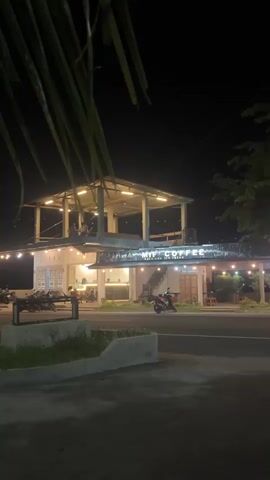 A night time shot of a multi-story coffee shop. The building is lit by string lights and interior lighting. There are several motorcycles parked outside. A few people are visible in the upper story and on the lower level. The street in front of the coffee shop is visible with a few more motorcycles passing by.