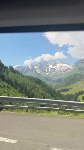 The video shows a view from inside a vehicle, looking out through a window at a mountainous landscape. The scene is dominated by lush green hills covered in dense pine forests, with a winding road in the foreground. In the distance, majestic snow-capped mountains rise against a bright blue sky dotted with fluffy white clouds. The sun appears to be shining, casting shadows on the mountainside and highlighting the vibrant greenery. The vehicle is in motion, as evidenced by the slight blur of the trees and the continuous movement of the scenery. A metal guardrail is visible along the edge of the road. The video is short and appears to be a scenic drive through the mountains.