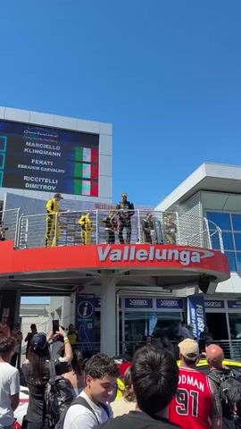 On a sunny day, confetti rains down on a crowd celebrating on a podium. The podium, labeled “Vallelunga”, is a raised platform with a metal railing, and sits in front of a modern building with glass windows. Above the podium is a large electronic scoreboard displaying results and the Italian flag. On the podium, several people in racing suits pop bottles and cheer. The crowd below, mostly young adults, holds up phones to capture the moment. They stand in front of sponsor signs, including one for “Derny”. A yellow sports car is also visible. The atmosphere is celebratory and lively.