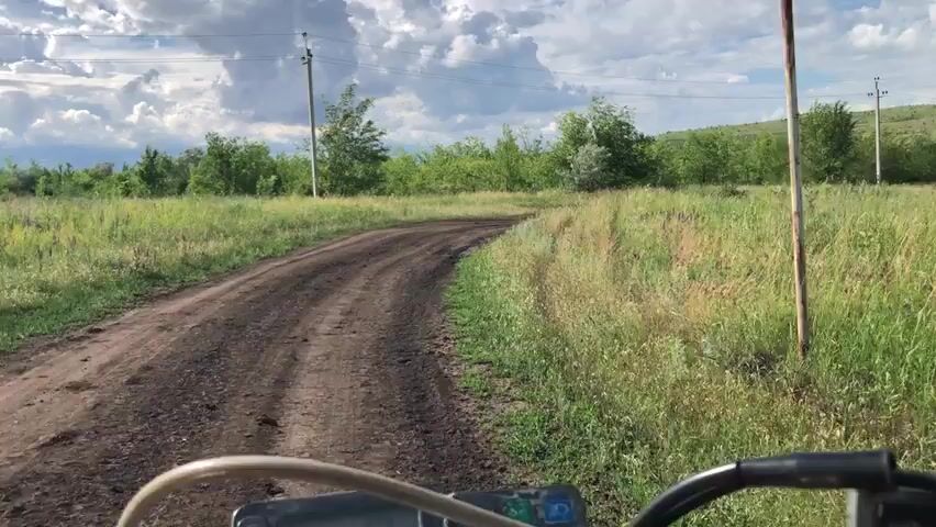 A first-person perspective video shows a dirt road winding through a grassy field. Trees line the sides of the road and a distant hill is visible with houses clustered on its slope. The sky is cloudy with patches of blue. Electrical poles and wires run alongside the road. The camera appears to be mounted on a moving vehicle, as indicated by the slight shakiness of the footage. The overall impression is of a rural landscape in a temperate climate.