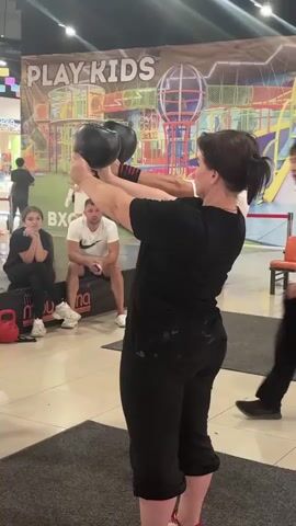 A woman in a black t-shirt and black pants is performing a kettlebell exercise. She is holding two black kettlebells in front of her face, with her arms bent. Her hair is pulled back in a ponytail. Behind her, a man in a white t-shirt and black pants is standing, looking at her. To the left of the man, a young person with dark hair is sitting on the floor, looking forward. Further in the background, there is a colorful play structure with the words "PLAY KIDS" visible on a banner. The scene appears to be indoors, possibly in a shopping mall or entertainment center, with a grey carpeted floor in the foreground. The overall atmosphere suggests a fitness demonstration or class.