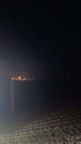 The video begins with a dark scene of a beach at night. In the distance, lights are visible, suggesting a town or resort. As the video progresses, fireworks begin to explode in the sky, illuminating the scene with bright flashes of light, primarily in red and orange hues. The fireworks create a dramatic effect against the dark sky. The foreground shows the sandy texture of the beach with some tire tracks or footprints. In the latter part of the video, a person wearing light-colored shorts is briefly visible on the left side of the frame, walking or standing on the beach and looking towards the fireworks. The environment appears to be an open beach area with the ocean likely being the source of the distant lights and fireworks display.
