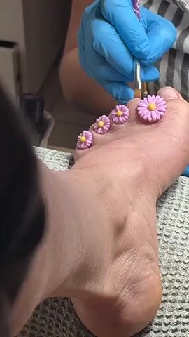 A close-up shot shows a person's foot with five small pink daisy-shaped charms attached to their toes. Another person, wearing blue nitrile gloves and a striped shirt, is using a tool to adjust the charms. The foot is resting on a grey textured towel. The background is blurred, but a black object and a white surface are partially visible.