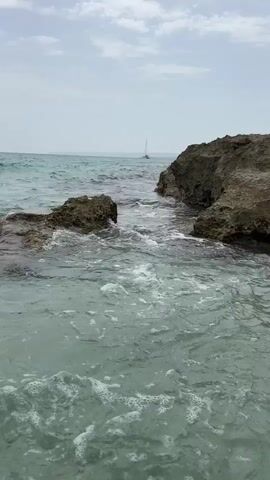 The video shows a view of the sea from a rocky coastline. The water is clear and turquoise, with waves gently lapping against the rocks. In the distance, a sailboat can be seen on the horizon. The sky is overcast, but the light is bright. The video captures the movement of the water, with splashes and foam as the waves break against the rocks. The overall impression is one of a peaceful and serene natural scene.