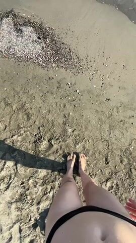 The video shows a first-person perspective of someone walking on a sandy beach. The person is wearing a black bikini bottom. Their bare feet are visible as they walk towards the ocean. The sand is light brown with some small pebbles and shell fragments scattered on it. The ocean water is clear with gentle waves. A shadow of the person is cast on the sand.