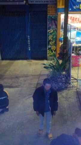 The video shows an outdoor street scene at night.  An adult man wearing a dark jacket, light pants, and sandals is standing on a tiled sidewalk.  A sign on a nearby business with the phone number "02633.840 532" is visible, along with other advertising. Another person is sitting on the ground, obscured by the standing man. Plants are placed next to a metal utility pole. The background shows a storefront with bright interior lights.