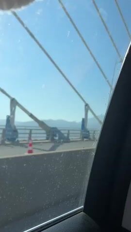 The video shows the interior of a car as it drives across a bridge. In the first shot, the view is through the passenger window, showing the bridge structure and the sky. The next three shots are from the perspective of the driver or from behind the driver's seat, showing the dashboard, steering wheel, and the road ahead. A person's head and shoulders are visible in the driver's seat. A GPS navigation system is mounted on the dashboard. The bridge appears to be a cable-stayed bridge with tall towers and numerous cables. The sky is clear and blue, and there is some water visible below the bridge. The overall impression is of a scenic drive across a large bridge on a sunny day.