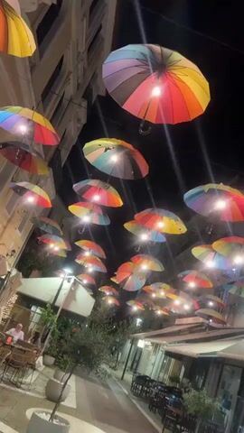 The video captures a night scene on a decorated street, likely a pedestrian walkway or commercial area. Numerous rainbow-colored umbrellas are suspended overhead, forming a vibrant canopy. Each umbrella is illuminated by a light, casting a bright glow and creating a festive atmosphere against the dark night sky.

On either side of the street, multi-story buildings house establishments, possibly cafes or restaurants, with outdoor seating areas. Tables and chairs are visible, and a few people can be seen sitting at them on the left side of the street. Potted plants and small trees line the pavement, adding to the ambiance. The street lights also contribute to the overall illumination. The scene depicts a lively evening setting in Karditsa, Greece.