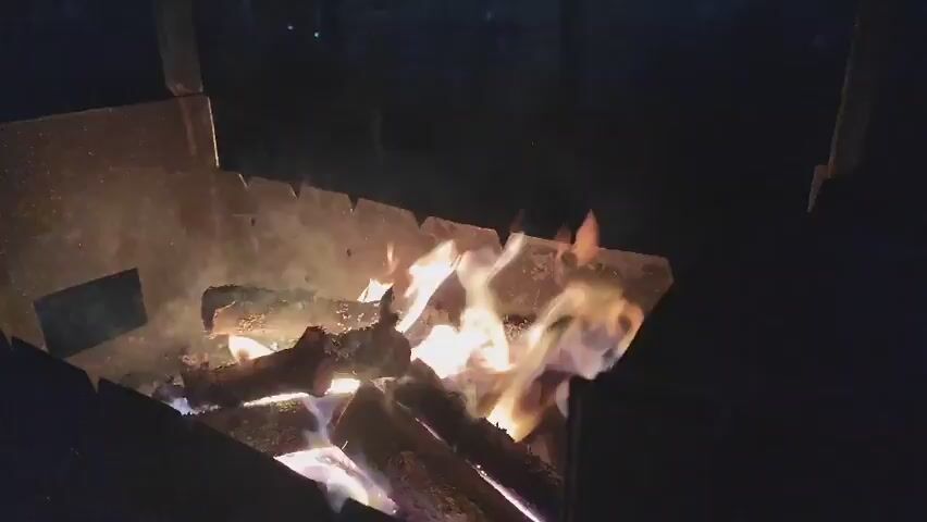 The video shows a close-up of a bonfire at night. Several logs are burning in a metal fire pit, emitting flames and smoke. The fire is bright and illuminates the surrounding area with a warm glow. The background is dark, suggesting it is nighttime. The fire pit appears to be outdoors. There are no people visible in the video, and no specific actions or interactions are occurring other than the burning of the wood.