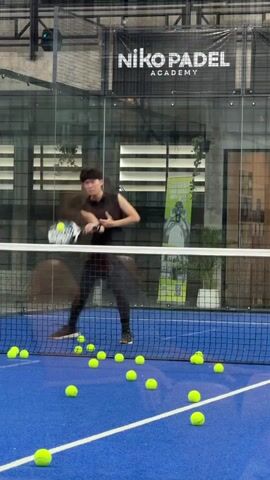 A young Asian man in athletic attire is on a blue padel court, holding a padel racket. He is in the process of hitting a yellow ball with his racket. Several yellow balls are scattered across the court. The court is enclosed by glass walls and a net separates the man from the camera. A sign above the glass wall reads "NIKO PADEL ACADEMY." Another sign in the background reads "DECATHLON." The man is mid-action, with one ball in the air near him, and another in motion from his racket.