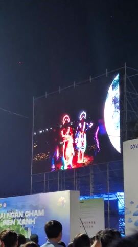 Here is a description of the video:

The video shows a large outdoor LED screen displaying three performers in traditional Vietnamese opera costumes. The performers are brightly lit with LED lights, creating vibrant colors and patterns on their costumes. They are standing on a stage with a red surface. The LED screen is set up outdoors at night, with a crowd of people watching the performance. The foreground shows the back of people's heads, implying they are part of the audience. A banner with text in Vietnamese is also visible in the foreground. The overall atmosphere suggests a cultural performance or festival.