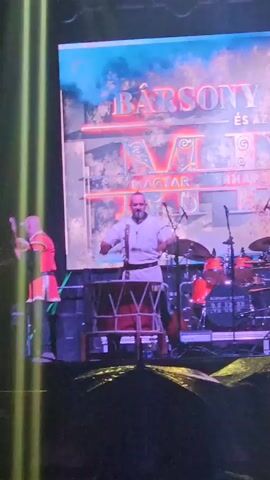 The video captures a live music performance on a stage. In the foreground, a man with a bald head and beard is playing a large drum with drumsticks. He is wearing a white tunic with suspenders and dark pants. To his left, another person in a red and yellow outfit is also on stage. Behind the drummer, a drum kit is set up, and a large screen displays text that appears to be a band name and other details. The stage is lit with bright lights, and the audience is visible in the lower part of the frame. The overall atmosphere suggests a dynamic and energetic performance.