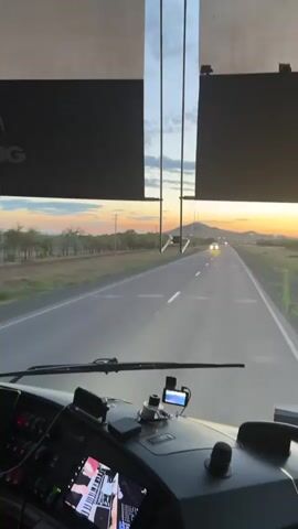 The video shows a view from the driver's seat of a vehicle, likely a bus or large truck. The perspective is looking forward through the windshield. The road stretches ahead, leading towards a distant hill. The sun is setting, casting warm hues of orange and yellow across the sky. A few clouds are visible. There are trees and fields on either side of the road. In the distance, other vehicles can be seen on the road, with their headlights on. The dashboard of the vehicle is visible in the foreground, with various buttons and screens. One screen displays what appears to be a piano keyboard. A phone or GPS device is mounted on the dashboard. The overall scene conveys a sense of travel and a scenic sunset.