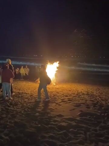 A group of people are gathered on a sandy beach at night around a large bonfire. The fire is burning brightly, casting a warm glow on the sand and the faces of those nearby. The ocean is visible in the background, with waves gently rolling onto the shore. The sky is dark, with a few distant lights suggesting a town or settlement. Most of the people are standing, watching the fire, while one person in the foreground is turned away from the camera, appearing to be interacting with the flames. Some individuals are wearing hooded jackets, suggesting a cool evening. The atmosphere appears relaxed and communal, with the bonfire serving as a focal point for the gathering.