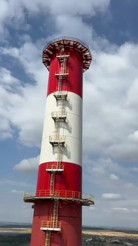 The video shows a close-up, upward-facing view of a tall, red and white striped industrial smokestack against a partly cloudy sky. The smokestack has a metal ladder with yellow platforms and railings running vertically up its exterior. The camera slowly zooms out, revealing more of the surrounding industrial landscape, including buildings, tanks, and what appear to be power lines. The perspective suggests the camera is also moving upwards or the smokestack is tilting. There are no people visible in the video.