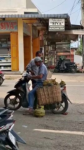 Two men are near a motorcycle. One man, wearing a blue shirt, jeans, and a white cap, is sitting on the motorcycle. He is holding the handlebars. A large woven basket is attached to the back of the motorcycle, containing several durians. A green gas tank is on the ground near the motorcycle. The second man, wearing a grey and blue checkered shirt and dark pants, is standing next to the first man, appearing to be talking to him. They are in front of a building with signs. One sign says "SIVA" and another says "Toko obat SIVA" and "NURZAHRI.SKM". There are other motorcycles and a building in the background. The ground is paved.