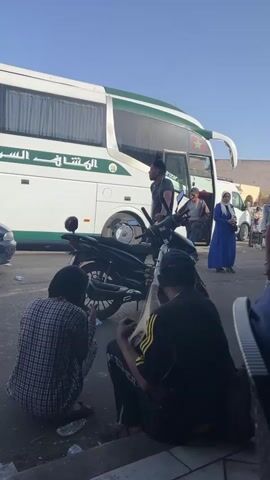 The scene captures a bustling outdoor environment near a large white and green bus, possibly a transport or tour vehicle given the Arabic writing on its side. Several people are visible around the bus, some boarding and others walking by, suggesting a moment of activity. To the immediate foreground, two people are seated on the curb, while a man stands nearby holding onto a black scooter. Litter is scattered on the ground around the seated individuals, hinting at a transient or frequently used space. The clear sky suggests a bright day.