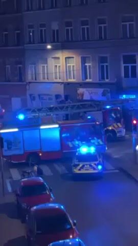 Here is a description of the video:

The video shows a street scene at night, likely in an urban setting. The primary focus is on emergency vehicles: several fire trucks and at least one police car. They are parked on the street with their blue emergency lights flashing. The buildings across the street are multi-story, with multiple windows visible. A street sign can also be identified above a shop with a sign that is illegible. A row of parked cars lines the left side of the street, closest to the camera. The perspective is from an elevated position, looking down at the street.