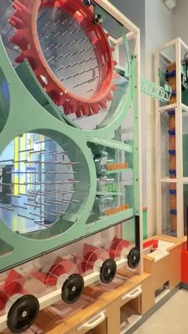 A close-up shot of a large interactive exhibit at a science museum focuses on a section designed to demonstrate energy generation. The exhibit features a large, clear cylinder filled with horizontal rods, creating a textured surface. Above this cylinder, a large red gear with protruding teeth is visible, suggesting a mechanism for storing or transferring energy. The entire structure is encased in a light green frame with visible bolts and connectors. To the right of the cylinder, text on the green frame reads "HELP POWER THE CLOCK SQUEEZE MORE ORANGES." Below the cylinder, a series of transparent plastic compartments hold what appear to be oranges. Each compartment has a red fan-like apparatus with black wheels at its base, hinting at a process of dispensing or manipulating the oranges. The lighting suggests an indoor environment, likely well-lit with ambient museum lighting. The camera is static, capturing the mechanical elements of the exhibit.