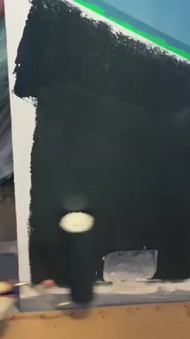 A person is painting a wall with a paint roller. The wall is mostly black, with some white showing on the left edge and at the bottom near a rectangular opening. The paint roller is dark colored and the handle is visible. The person's hand is gloved and holding the roller. The background is blurry and shows a green and blue area at the top. The floor is made of cardboard or wood.