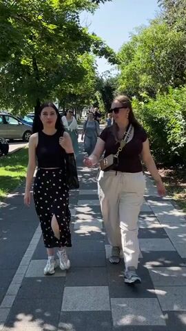 Two women walk side-by-side on a sidewalk lined with trees. The woman on the left wears a black sleeveless top and a black skirt with white polka dots, carrying a black bag in her left hand. The woman on the right wears a brown t-shirt, light-colored pants, sunglasses, and a small crossbody bag. Several other people walk behind them on the same sidewalk, and cars are parked on the left side of the street. The scene is outdoors on a sunny day.