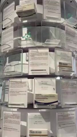 The video shows a close-up of a rotating display stand filled with medication boxes and printed materials. The stand is cylindrical and tiered, made of clear acrylic or plastic. Each level of the stand holds various items, including medication boxes with labels in both English and Chinese, along with informational sheets or pamphlets. The background is blurred, suggesting an indoor environment with some foot traffic. The overall lighting is bright. The stand rotates slowly, allowing views of the different products and informational materials.