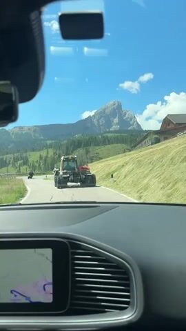 The video shows a view from inside a car, looking forward through the windshield. The car is on a paved road that winds through a mountainous, green landscape. In the distance, a large, rugged mountain peak dominates the skyline. The road is flanked by grassy slopes and scattered trees, with a few buildings visible on the right side.

In the middle ground, a red and white tractor is driving in the same direction as the car, on the right side of the road. The tractor appears to be a heavy-duty vehicle, possibly used for agricultural or construction purposes. A smaller, dark-colored vehicle is visible further ahead of the tractor.

The sky is a bright blue with scattered white clouds. The sun is shining, casting a bright light on the scene.

In the foreground, the dashboard of the car is visible, including a large touchscreen display showing a navigation map. The video is shot from the perspective of the driver. The overall impression is that of a scenic drive through a rural, mountainous area.