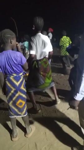 At night, an outdoor gathering features a group of people, mostly viewed from behind. A young girl in a purple shirt and patterned skirt stands to the left, looking towards a woman in a white shirt and similar skirt. Behind them, other figures dance and move in the dimly lit space. A man stands partially in the foreground, to the right. The ground appears to be sandy or dusty. A person films the event. Music is audible.