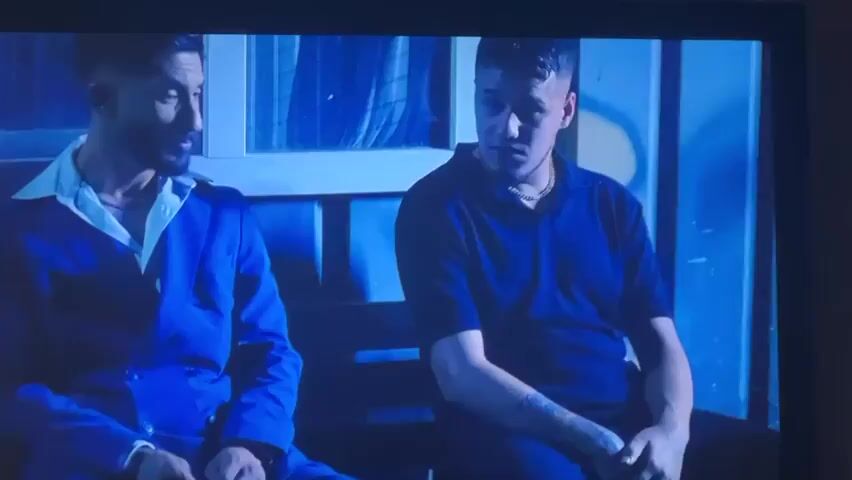 The video shows two men sitting on a bench in a room with blue lighting. The man on the left is wearing a blue jacket with a white collar and a silver chain. The man on the right is wearing a blue short-sleeved polo shirt and a silver chain. They are sitting in front of a wall with a window that has a blue curtain. The wall also has some graffiti on it. The man on the right is speaking.