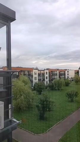 The video shows a view from a balcony or window overlooking a courtyard. In the foreground, a railing is visible on the left side. The courtyard is covered with green grass and has several trees of varying sizes and shapes scattered throughout. A paved pathway runs diagonally across the courtyard from the bottom right towards the center. In the background, there are modern apartment buildings with balconies and windows. The sky is overcast with clouds. The overall scene depicts a residential area with a well-maintained green space.