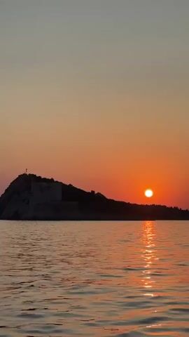 The video displays a wide shot of a tranquil ocean at sunset. The sun, a bright orange orb, is partially submerged behind a dark, rocky island that features what appears to be a stone structure on its peak. The sky is a gradient of warm oranges and softer yellows, transitioning to a pale blue at the top of the frame. The water reflects the vibrant colors of the sunset, with shimmering ripples across its surface. The overall scene evokes a sense of peace and natural beauty.