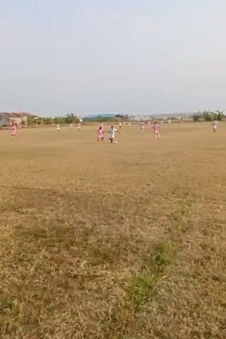 A soccer game is being played on a dry, grassy field under a clear sky. Several players, mostly wearing pink jerseys, are scattered across the field. One player in a pink jersey is dribbling a soccer ball, while another player in a white jersey is attempting to tackle them. Another player in a pink jersey is falling to the ground. In the background, there are some buildings and trees. The overall impression is of a casual soccer match in a rural or undeveloped area.