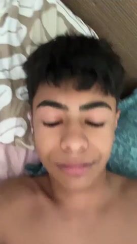 The video is a close-up, point-of-view shot of a young person lying in bed. The person has dark, short hair and dark eyebrows. They are looking at the camera and making facial expressions and hand gestures. The background consists of bedding with a pattern of brown and white shapes, and a pillow with a blue floral pattern. The lighting appears to be natural. The person touches their hair and mouth, and then waves at the camera.