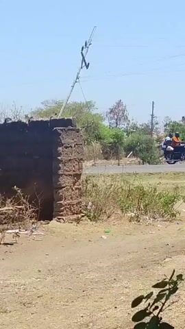 A utility pole leans precariously at an angle, its wires dangling. To the left, a crumbling brick wall stands partially intact. The ground is dry and dusty, with patches of scrubby vegetation and scattered litter. In the background, a road curves away, and a motorcycle carrying two individuals travels along it. The sky is clear and blue, suggesting a sunny day.