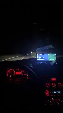 The video shows the interior of a car driving at night on a road. The dashboard instruments are illuminated in red. A GPS navigation screen is mounted on the dashboard, displaying a map. The road ahead is dark, with headlights of other vehicles visible in the distance. The driver's hands are not visible.