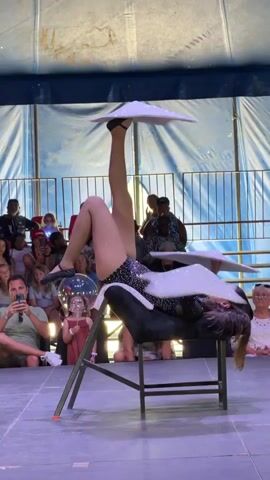 A young woman with long brown hair, wearing a sparkly black leotard and fishnet stockings, performs a balancing act in what appears to be a tent or circus-like venue. She is positioned on a black cushioned stool that is attached to a metal chair. Her legs are extended upwards, and she is balancing two white, sparkly plates on the soles of her feet. Another plate is balanced on her forehead, and a fourth plate is positioned on her chest. Her body is contorted in a difficult yoga-like pose. In the background, an audience of people, including children, watch the performance. The environment is dimly lit, with a blue and white striped fabric backdrop.