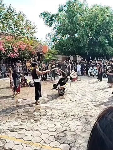 The video shows a group of people gathered in an outdoor space. Many are wearing black outfits and some appear to be participating in a performance or martial art demonstration, with at least one person falling to the ground. Some of the participants are using what appear to be rattan sticks. There's an audience watching the performance, including some children. The setting seems to be a courtyard or plaza with a tiled floor. There are trees and a building in the background. Some individuals are identified as "crew" on their shirts.