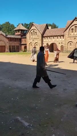 In an outdoor courtyard with wooden buildings in the background, five people are engaged in what appears to be a staged reenactment or performance. One person in the foreground is facing away from the camera, holding a sword. In the middle ground, four people are positioned. Two men, one in a light-colored robe and the other in armor, stand facing the person in the foreground. Between them, two women are dressed in traditional clothing, one in a red and orange dress and the other in a brown and gold dress. To the right, a person in dark clothing with a cape is also present. The ground is dirt and gravel, and the sky is clear blue. The overall scene suggests a historical or fantasy setting.