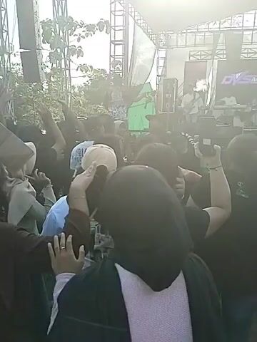 The video shows a live outdoor concert. The stage is equipped with a large screen displaying the logo "DVS Mediatama". A band is performing on stage, with a female singer taking center stage. In front of the stage, a crowd of people are watching and dancing. Some people are holding up their phones to record the performance. Behind the crowd, there is a tent where people are standing and watching the concert. A cameraman with a professional camera is recording the performance from behind the tent. The environment appears to be open and potentially rural, with trees visible on the edges and a glimpse of a field in the distance.