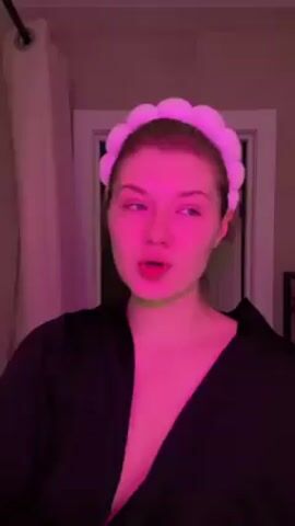 A woman with fair skin is centered in a low-angle shot against a bathroom background with a closed white door.  The woman has a light pink puffy headband on, blue eyes, and is wearing a black robe that is slightly open, revealing her chest area. She leans her head on her left hand, as if tired, and opens her mouth, appearing to sing or speak along with music. The lighting is primarily pink, giving the scene a pink hue, with a slight green tinge on her face. There is a white shower curtain on the left.