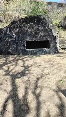 A video shows a large, dark rock formation with a rectangular opening carved into its face. The opening is dark and appears to be a cave or shelter. The rock is surrounded by dry, cracked earth and sparse vegetation. In the background, there are trees and hills. Towards the end of the video, a small, open-air pavilion with a tiled roof is visible. Two people are sitting on a bench under the pavilion. One person is wearing a red and white jacket and a headscarf, while the other is wearing dark clothing. There are also two helmets visible near the bench. Shadows of trees are cast across the ground, suggesting it is daytime.