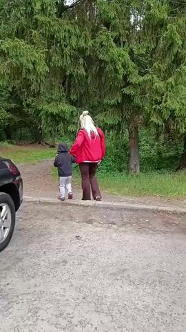 The video shows a family in Staryi Pokrov, Russia, preparing for an outdoor activity. In the foreground, a black SUV is parked on a gravel surface. Two people, a woman and a young child, are walking away from the camera towards a wooded area with tall evergreen trees. The woman has blonde hair and is wearing a red jacket and dark pants. The child is wearing a dark hooded jacket and gray pants. The SUV's rear hatch is open, and a person wearing a denim shirt and black pants is seen reaching into the cargo area, which contains several large plastic bottles of water. The scene appears to be taking place during the daytime, with overcast skies. The overall impression is one of casual preparation for an outing in a natural setting.