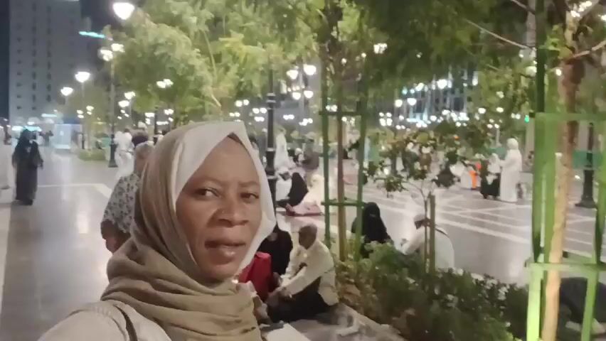 The video shows a woman wearing a tan hijab standing in a public area at night. She is facing the camera and looking at the lens. Behind her is a square paved area with people sitting and walking around. Some of the people are wearing traditional Islamic clothing. There are small trees planted around the square, and streetlights illuminate the area. The camera is held at about chest level, and the video is slightly shaky.
