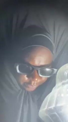 A dark-skinned person wearing a black head covering and black-rimmed glasses looks into the camera, illuminated by a lamp. They say "You can call me the Queen Mother" and then laugh. The background is dark.